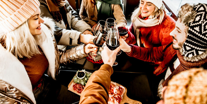 Group of smiling friends drinking and toasting red wine at bar restaurant in winter time - Friendship concept with young happy people having fun together at xmas happy hour - Focus on glasses