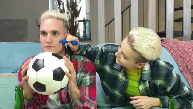 Young stylish white couple preparing to cheer France soccer team on championship at home. Happy young woman painting French flag on male face indoors. Support France football at world cup