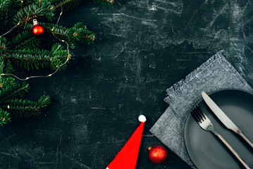 Table setting with black plate, fork, knife, santa hat, fir branch and baubles on black background with copy space. Christmas concept. Top view, flat lay