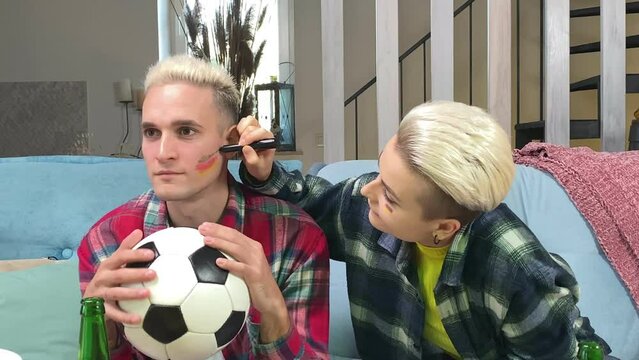 Young stylish white couple preparing to cheer Germany soccer team on championship at home. Happy young woman painting German flag on male face indoors. Support Germany football at world cup