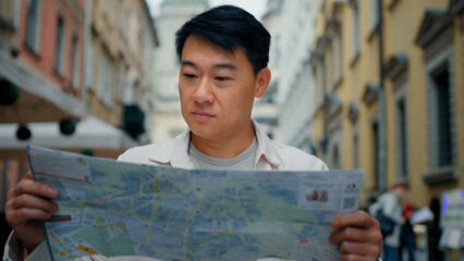 Asian male tourist traveling in new city standing outdoors looking at paper map in search of location using travel guide to get directions young man traveler international student searching right way