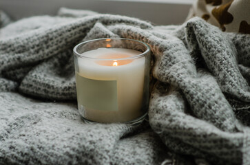 A candle in a glass on a windowsill on a gray blanket. Cozy autumn atmosphere.