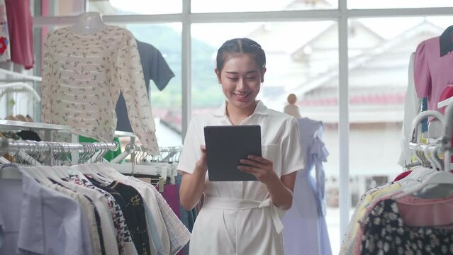 Asian Female Owner Of Clothing Store Using A Tablet Then Smiling To The Camera While Checking Stock On Rails
