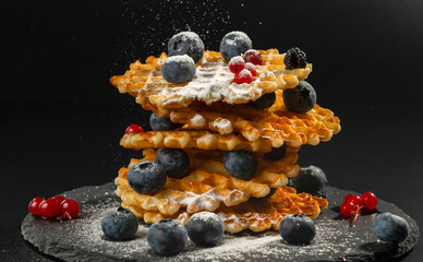 Fresh crispy Belgian waffles with fresh blueberries blackberries cranberries and red currants sprinkled with powdered sugar on a black stone plate on a black background