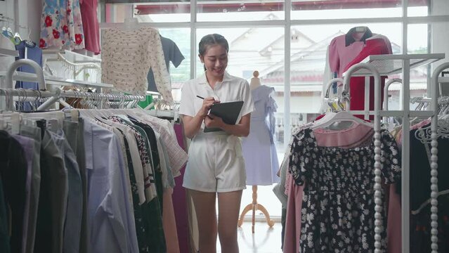 Asian Female Owner Of Clothing Store Taking Note On The Tablet And Smiling To The Camera While Walking Around Checking Stock On Rails
