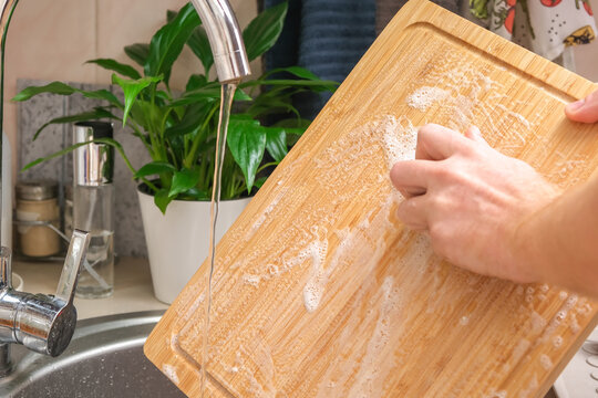 A man washes a wooden bamboo cutting board in the kitchen sink under running water. Gentle hand washing of a wooden cutting board. Cleaning dirty kitchen wood products.