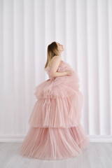 A girl in a beautiful lush long dress of the color of a dusty rose stands near a light wall....