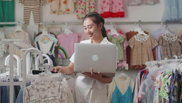 Asian Female Owner Of Kids Clothing Store Enjoys Using A Laptop To Check Stock On Rails
