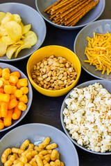 Salty snacks assortment shot on dark slate table. The picture includes potato chips, popcorn, peanut, cheese sticks, ketchup sticks, tzatziki chips, smoky flips, and others