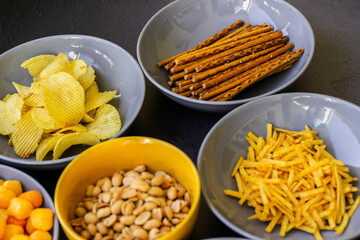 Salty snacks assortment shot on dark slate table. The picture includes potato chips, popcorn, peanut, cheese sticks, ketchup sticks, tzatziki chips, smoky flips, and others