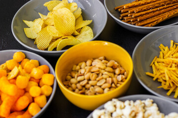 Salty snacks assortment shot on dark slate table. The picture includes potato chips, popcorn, peanut, cheese sticks, ketchup sticks, tzatziki chips, smoky flips, and others