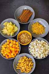 Salty snacks assortment shot on dark slate table. The picture includes potato chips, popcorn, peanut, cheese sticks, ketchup sticks, tzatziki chips, smoky flips, and others