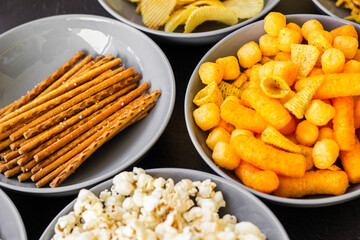 Salty snacks assortment shot on dark slate table. The picture includes potato chips, popcorn, peanut, cheese sticks, ketchup sticks, tzatziki chips, smoky flips, and others