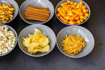 Salty snacks assortment shot on dark slate table. The picture includes potato chips, popcorn, peanut, cheese sticks, ketchup sticks, tzatziki chips, smoky flips, and others