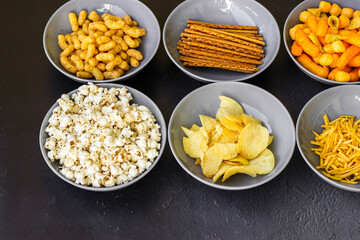 Salty snacks assortment shot on dark slate table. The picture includes potato chips, popcorn, peanut, cheese sticks, ketchup sticks, tzatziki chips, smoky flips, and others