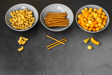 Salty snacks assortment shot on dark slate table. The picture includes potato chips, popcorn, peanut, cheese sticks, ketchup sticks, tzatziki chips, smoky flips, and others