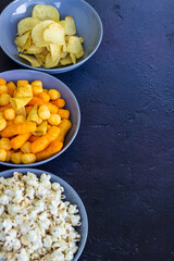 Salty snacks assortment shot on dark slate table. The picture includes potato chips, popcorn, peanut, cheese sticks, ketchup sticks, tzatziki chips, smoky flips, and others