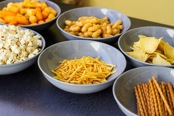 Salty snacks assortment shot on dark slate table. The picture includes potato chips, popcorn, peanut, cheese sticks, ketchup sticks, tzatziki chips, smoky flips, and others