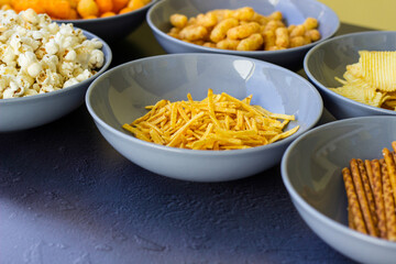 Salty snacks assortment shot on dark slate table. The picture includes potato chips, popcorn, peanut, cheese sticks, ketchup sticks, tzatziki chips, smoky flips, and others
