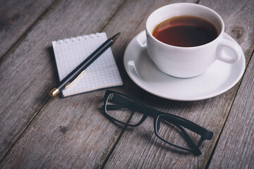 Opened notebook, pen, glasses, and coffee cup on wood table with blank area in morning time with vintage filter effect