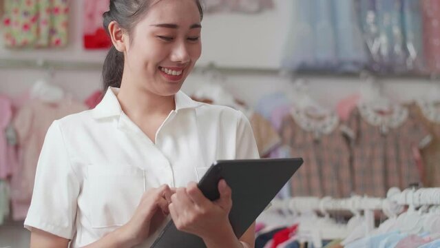 Close Up Of Asian Female Owner Of Kids Clothing Store Using A Tablet Then Smiling To The Camera While Checking Stock On Rails
