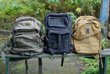 a row of three large army green black backpacks stand on a wooden bench in the street