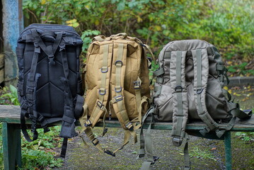 a row of three large army green black backpacks stand on a wooden bench in the street