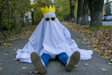 Holiday Halloween. Sitting on the sidewalk is a child dressed as a ghost made of a white sheet and...