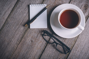 Opened notebook, pen, glasses, and coffee cup on wood table with blank area in morning time with vintage filter effect