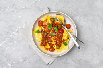 Traditional Italian food pasta ravioli with tomato sauce and olives with basil and cheese in a beige plate on a gray background.
