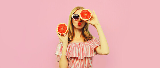 Summer portrait of beautiful young woman with slice of juicy grapefruit blowing her lips sending sweet air kiss wearing heart shaped sunglasses on pink background