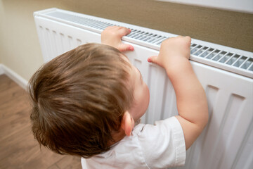 Toddler baby holding on to the radiator, child hand on the heating system close-up. White radiator for heating the home room and children's hand