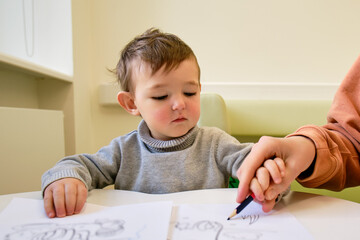 Mother teaches to draw toddler baby with a pencil on a white sheet of paper. Parent with child boy learning to color coloring book. Kid aged one year and three months