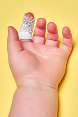 Baby s hand with a bandaged finger on a yellow studio background, close-up. Injured index finger of a child wrapped in a white bandage. Kid boy aged one year and three months