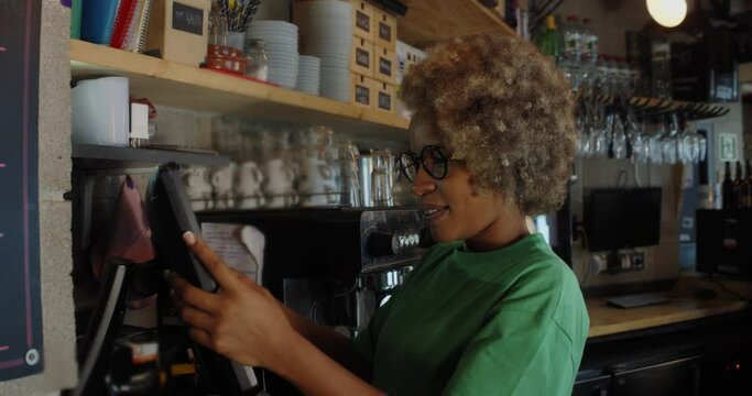 Happy African American female waitress using cash register while reworking in a pub.