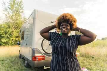 Body positive and independent Black woman looking at camera, holding her hands in her afro hairstyle hair, and standing in front of her white van. Outdoor shot. High quality photo - Powered by Adobe