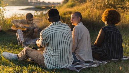 Lazy warm summer or autumn evening by the river. Three multiracial friends sitting on checked picnic blanket and taking. Blurred caucasian guy with acoustic guitar in the background. High quality
