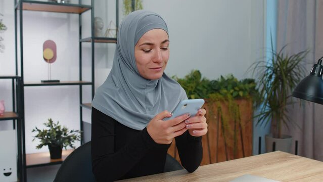 Young Muslim Business Woman Wearing Traditional Hijab Headscarf In Modern Home Office Workspace Texting Share Messages Content On Smartphone Social Media Applications Online, Watching Relax Movie