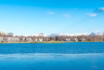 Westchester Lagoon with small island on the lake and row of duplex houses with downtown Anchorage buildings office towers in background