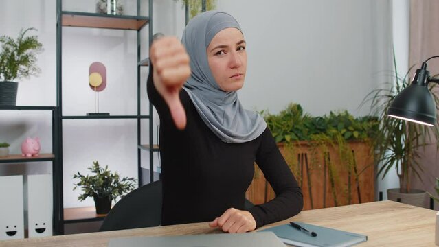 Dislike. Upset young muslim business woman wearing hijab headscarf showing thumbs down sign gesture, expressing discontent, disapproval, dissatisfied bad work. Displeased girl at home office workspace