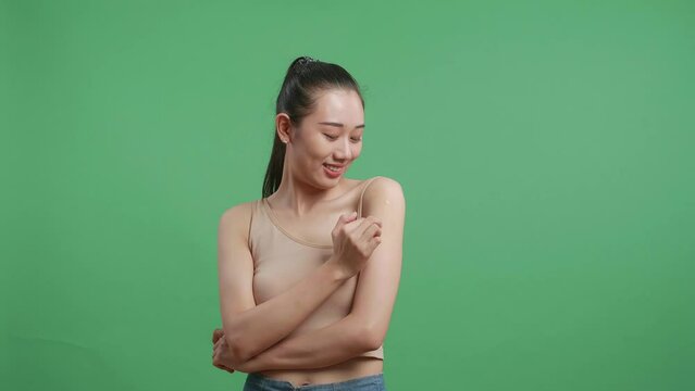 Asian Woman Applying Body Lotion To Her Shoulder And Smiling While Standing On Green Screen Background In The Studio
