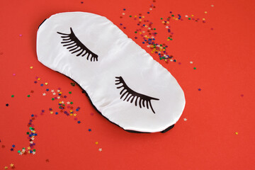 sleeping mask with painted eyelashes and glittery star-shaped confetti on red background dream concept