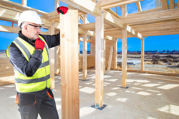 Fototapeta premium Builder man thought. Builder with board in his hand. Man is loader for construction company. Builder is standing in yellow vest scratching his chin. Guy builds house out of wood. Wooden house frame