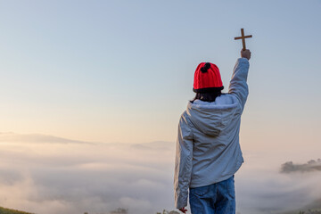 Human holding christian cross Prayer to God on mountain top sunrise foggy background.christian...