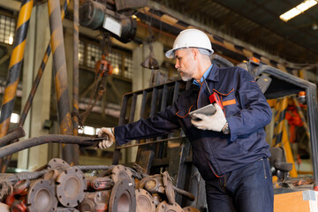 A machine factory engineer is inspecting the machines in the factory with a tablet or laptop and with a deliberate eye check. The factory is big and old.