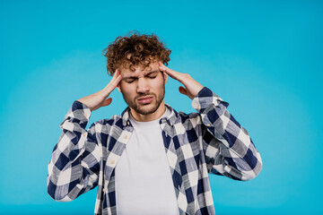 Young attractive man in printed shirt feels anxious suffering from headache holds his head, isolated blue background