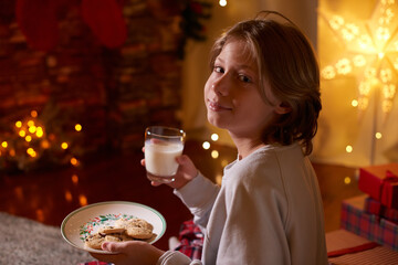 Caucasian fair hair boy waiting for Santa holding in hands glass of milk, cookies sitting on floor at home in Christmas Eve. Happy holidays time Winter spirit Getting cozy vibes Christmas decoration