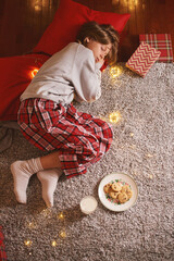 Cute child waiting for Santa holding in hands glass of milk and cookies sitting on floor at home in Christmas. Candid moment Winter time season Dreamy look about miracle
