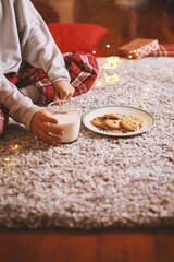 Cute child waiting for Santa holding in hands glass of milk and cookies sitting on floor at home in Christmas Eve. Christmas decoration Candid moment Winter time season Dreamy look about miracle