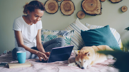 Happy young woman freelancer is working with laptop typing text sitting on bed at home with cute pedigree dog lying near her. Distant online work and people concept.